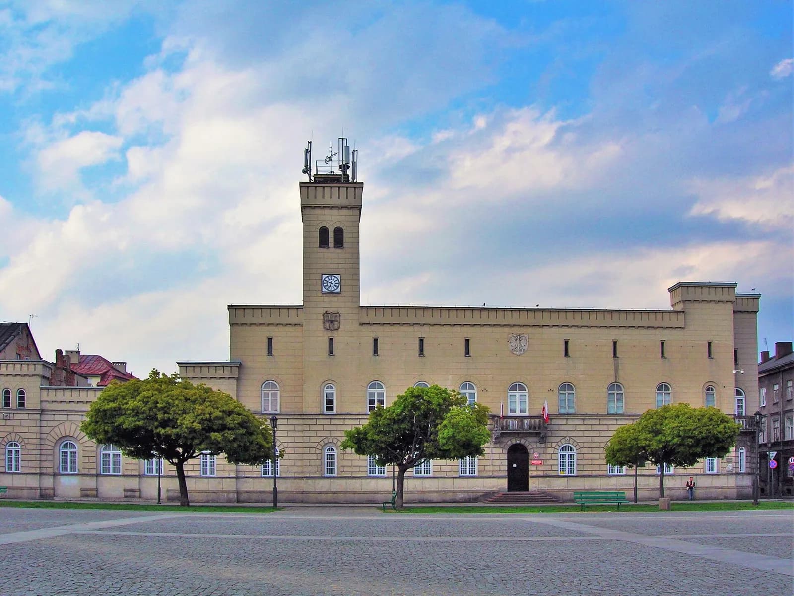 A historic building with a large central tower and arched windows, surrounded by green trees, under a blue sky with white clouds.