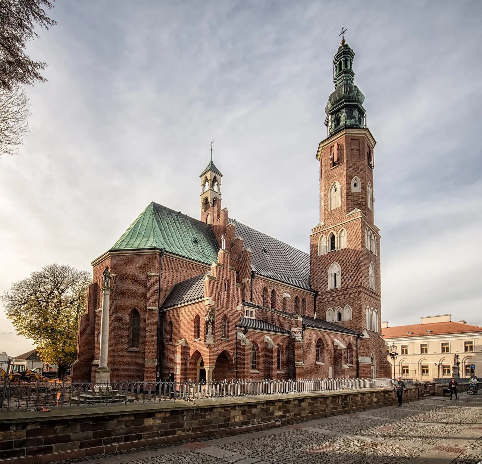 A large red brick church with a green roof stands prominently under a partly cloudy sky, featuring two towers with spires, one of which is significantly taller and intricately designed.